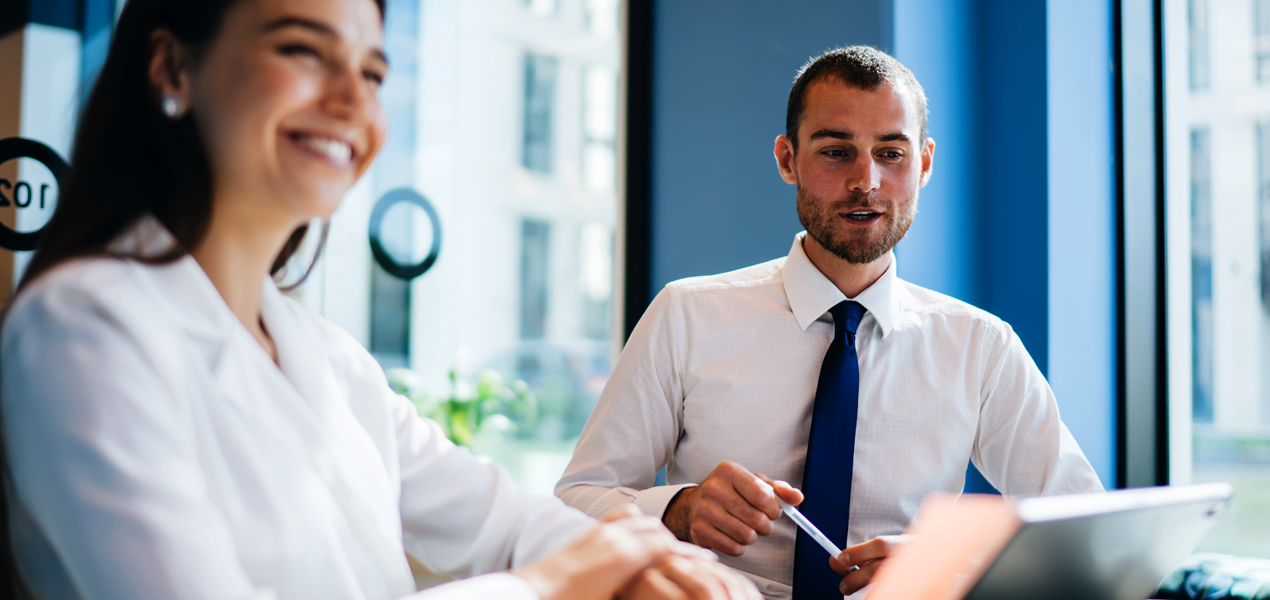 Deux personnes en tenue professionnelle sont assises à une table dans un bureau moderne avec de grandes fenêtres.