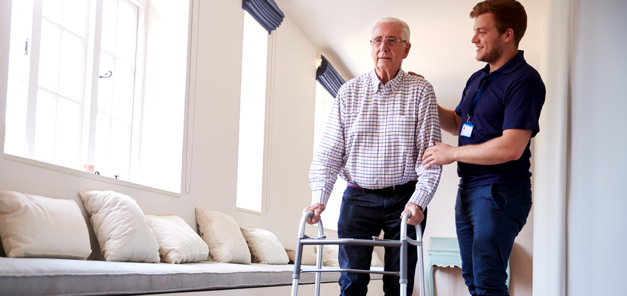 A healthcare worker in uniform assisting an elderly man to walk.