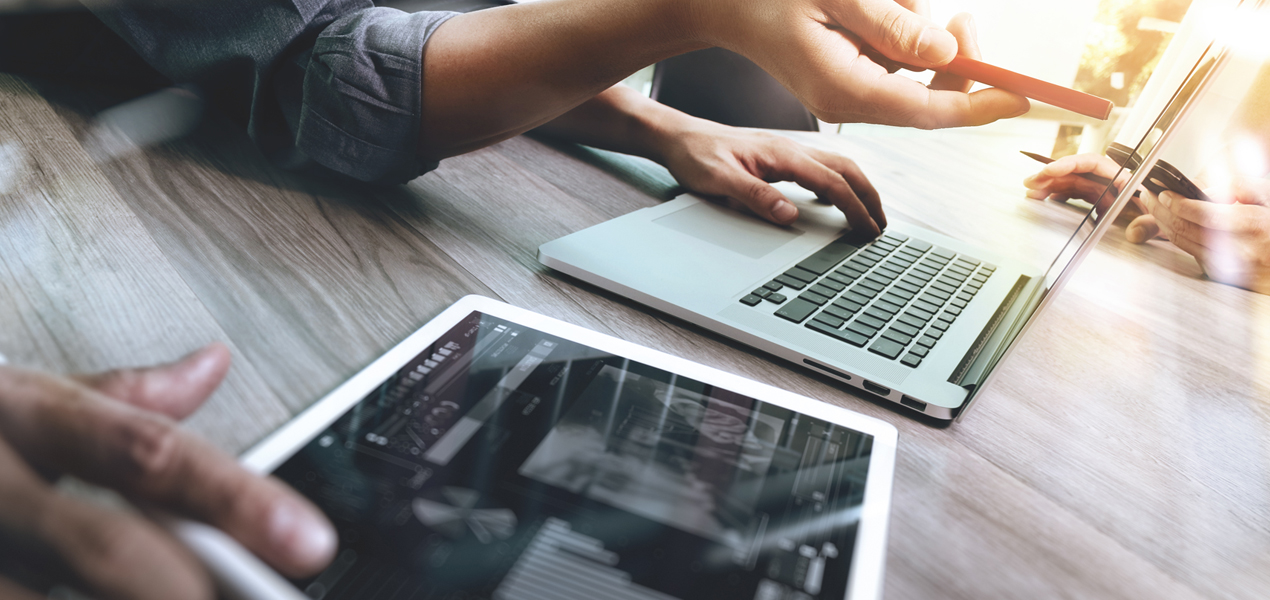 Close up of the hands of two people working side-by-side, one working on a tablet and one on a laptop