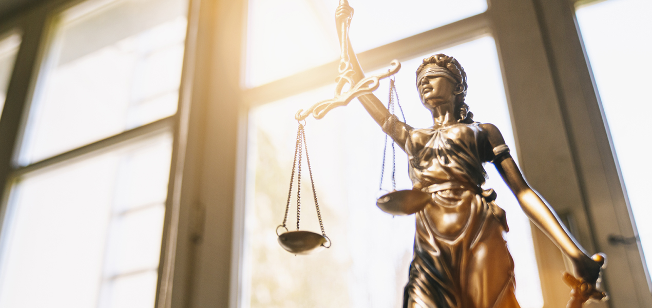 Une statue en bronze de la Justice tenant une balance et une épée se tient devant une grande fenêtre éclairée par le soleil, symbolisant l’équité et la loi. La figure est les yeux bandés